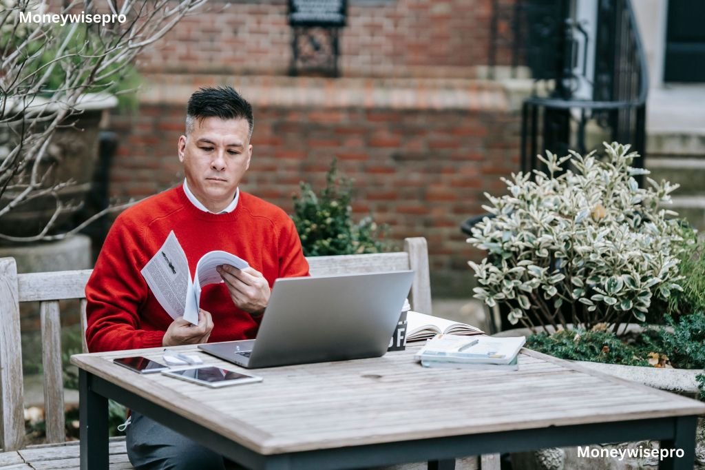 A person sits at a house garden table reviewing paperwork while checking a laptop screen .