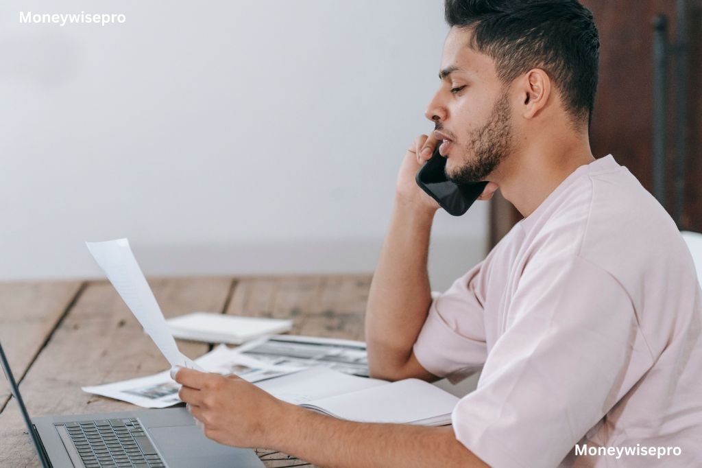 A person at a kitchen table talks on the phone while reviewing a paper bill.