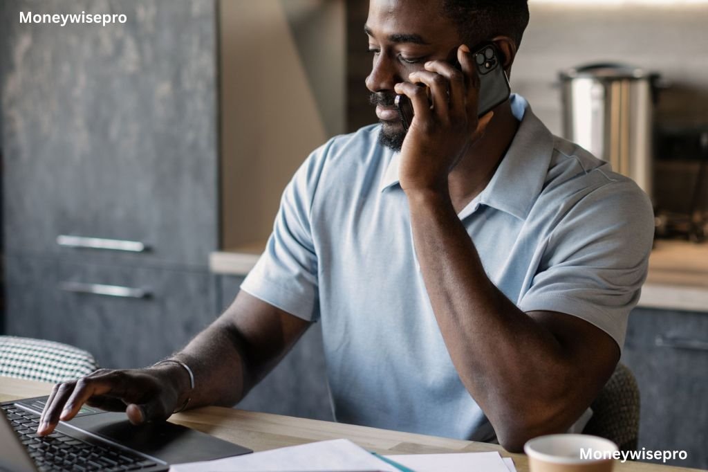 Man calmly talking on phone while taking notes on laptop in kitchen