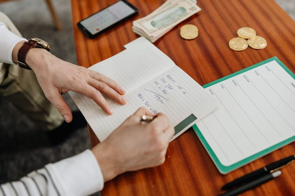 A hand writing numbers in a notebook next to a calculator